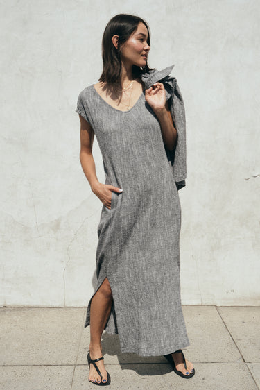 Woman standing on the sidewalk, outside wearing a salt and pepper yarn dye long sheath dress with a cap sleeve. She is holding a matching jacket over her shoulder.