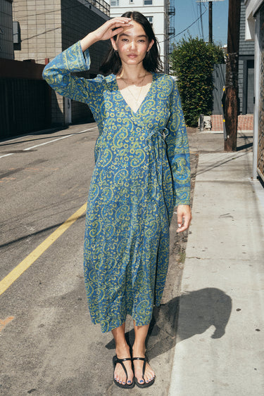 A woman standing in the street wearing a long sleeve blue paisley tapestry midi wrap dress.