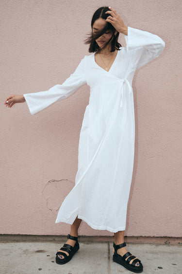 A model wearing a white maxi wrap dress with bell sleeves and an empire waist, standing against a pink background.