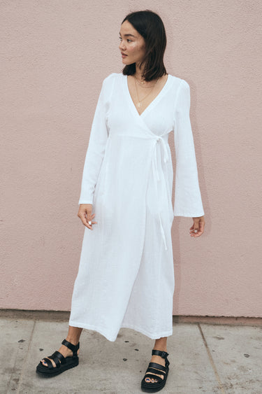 A model wearing a white maxi wrap dress with bell sleeves and an empire waist, standing against a pink background.