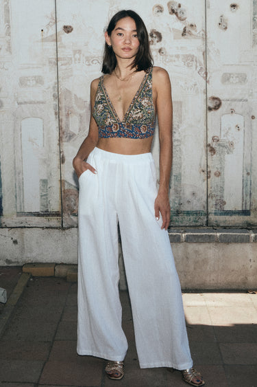 A woman standing outside by a mural wearing white full leg trousers with a tapestry bra top.