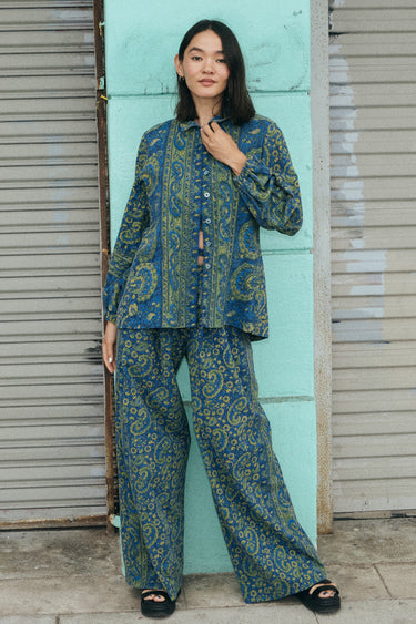 A woman standing outside by a wall wearing a shirt jacket cut from a dark blue paisley tapestry, with a matching trouser.