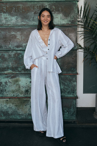 A woman standing outside by a verdigris wall wearing a white with black stripe gauze trouser with a matching jacket and bra top.