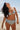 A woman on a beach wearing a white ground with lavender, blue and green floral tapestry bikini bottom with a high rise and matching bikini top.