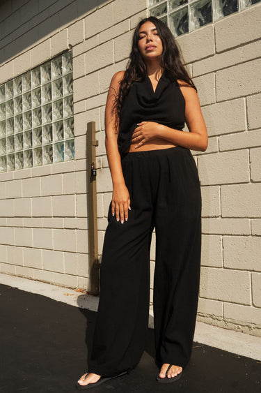 A woman standing outside next to an off white building wearing black trousers and a matching halter top.