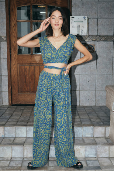 A woman wearing a blue crop top with a dark blue paisley printed design, paired with matching blue trousers, standing near a doorway.