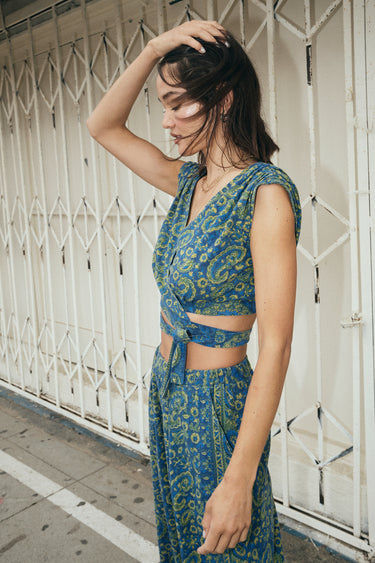 A woman wearing a blue crop top with a dark blue paisley printed design, paired with matching blue trousers, standing near a white security gate.