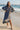 Woman in a grey dress standing on a beach with a pier in the background.