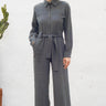 Woman wearing a gray jumpsuit standing against a white wall.