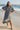 Woman in a long, light-colored dress standing on a beach with a pier in the background.