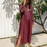 A woman wearing a long-sleeved, button-front burgundy maxi shirt dress with side slits, paired with beige ankle boots, standing outdoors in front of a white wooden fence and a leafy plant.