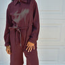 A woman wearing a long-sleeve maroon jumpsuit with a gray abstract graphic on the chest, standing in front of a white garage door.