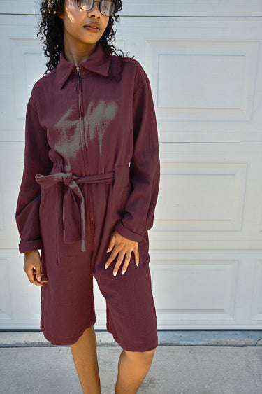 A woman wearing a long-sleeve maroon jumpsuit with a gray abstract graphic on the chest, standing in front of a white garage door.