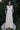 Woman wearing an alabaster dress standing on a white chair with greenery in the background