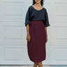 A woman stands in front of a white garage door wearing a black short-sleeve top and a high-waisted, knee-length burgundy skirt, paired with black Mary Jane shoes.