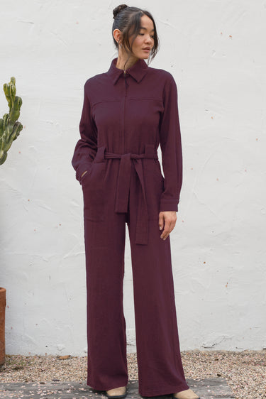 Woman wearing a black fig jumpsuit standing against a white wall.
