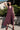 Woman wearing a burgundy dress standing outdoors with greenery and a building in the background