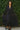 Person wearing a long black shirt and skirt astanding  against a green leafy background