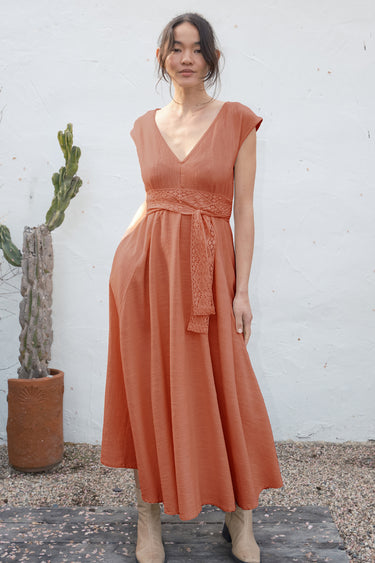 Woman wearing a rust-colored dress standing against a white wall with a cactus plant.