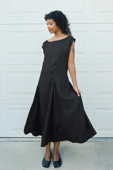 A woman wearing a black sleeveless midi dress with a boat neckline and a flared skirt, standing in front of a white garage door.