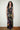 A woman with curly dark hair stands indoors wearing a sleeveless black maxi dress with a colorful celestial print of stars, moons, and suns. She is standing on a woven rug against a plain off-white wall.