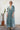 A woman stands outdoors against a white wall wearing a long-sleeve bohemian-style maxi dress with a blue, green, and white paisley print. She is wearing tan boots, with a potted cactus to her left.