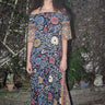 Woman standing in garden wearing a maxi dress with a bardot neckline, cut in a black ground sunflower tapestry. 