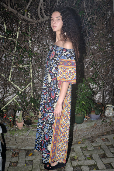 Woman standing in garden wearing a maxi dress with a bardot neckline, cut in a black ground sunflower tapestry. 