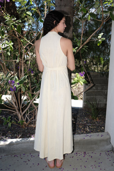 A woman outside wearing an alabaster off white maxi caftan with deep cut arm holes and bias cut sides, featuring an obi belt, back view.