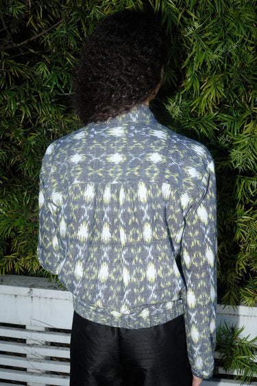 Woman outdoors wearing a gray with white and yellow ikat zip front jacket with black pants, back view.