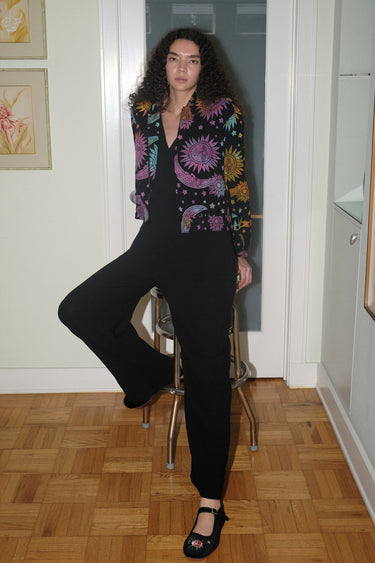 A woman wearing a cropped black ground with rainbow suns, moons and stars tapestry jacket with a zip front, standing in a home environment.