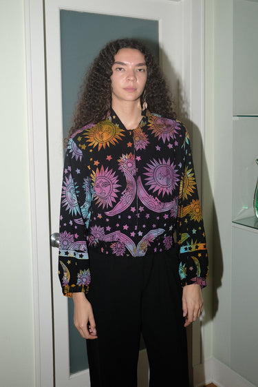A woman wearing a cropped black ground with rainbow suns, moons and stars tapestry jacket with a zip front, standing in a home environment.