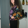 A woman wearing a cropped black ground with rainbow suns, moons and stars tapestry jacket with a zip front, standing in a home environment.