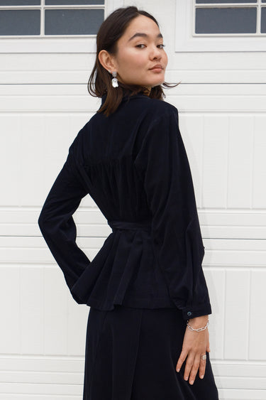 A woman wearing an ebony black  corduroy shirt with long sleeves and an obi belt standing by a white wall, back view.