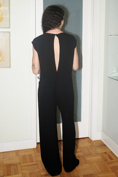 A woman standing in indoor setting wearing a slim black full length jumpsuit, back view.
