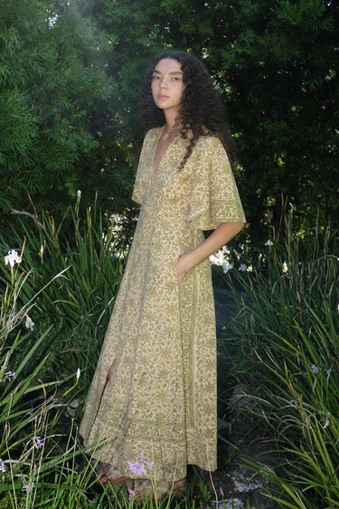 woman standing in garden wearing a light green print tapestry dress with zip front, v neck and elbow length sleeves.