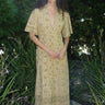 woman standing in garden wearing a light green print tapestry dress with zip front, v neck and elbow length sleeves