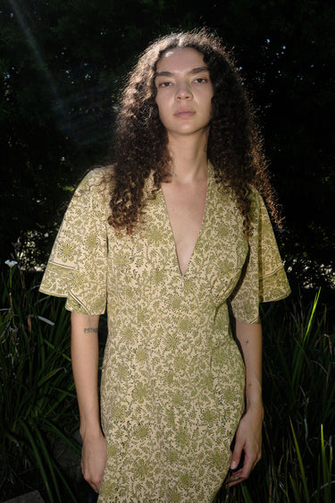 woman standing in garden wearing a light green print tapestry dress with zip front, v neck and elbow length sleeves