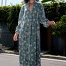 Woman wearing a long tarragon colored floral tapestry caftan standing outdoors with greenery in the background.