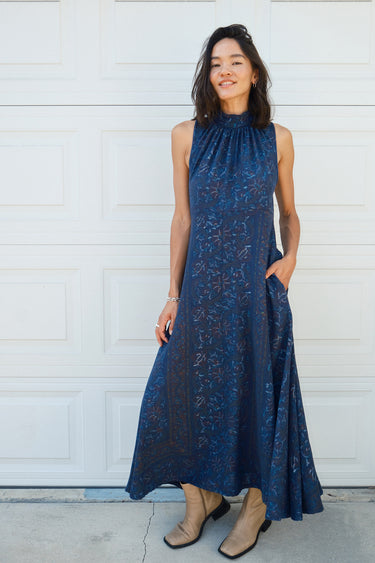 A woman standing in front of a door, wearing a navy dress with a band collar, empire waist, and hand woven, hand block printed Indian Tapestry fabric. She has a smiling expression and is wearing tan boots. 