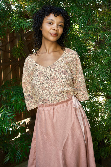 Woman wearing a tapestry top and pink skirt standing in front of green foliage