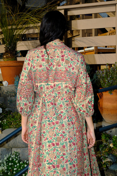 A woman standing outside wearing a beige ground with red and brown belted floral caftan. Back View 