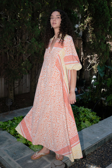 A woman standing in a garden wearing a pink and orange maxi dress with three-quarter sleeves and a belt, the dress is made of cotton hand block printed fabric.