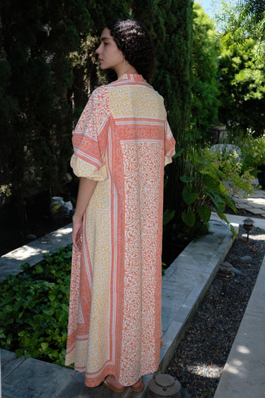 A woman standing in a garden wearing a pink and orange maxi dress with three-quarter sleeves and a belt, the dress is made of cotton hand block printed fabric.