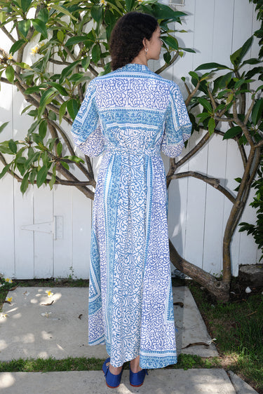 A woman standing on a pathway with a white and blue floral print maxi dress with three-quarter sleeves and a belt, wearing blue shoes, back view.