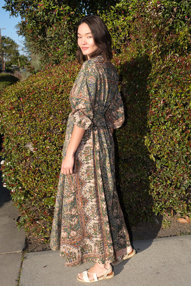 A woman wearing a maxi caftan with a floral pattern in a color that could be described as celadon green ground with tan, brown and blue, belted, standing outdoors.