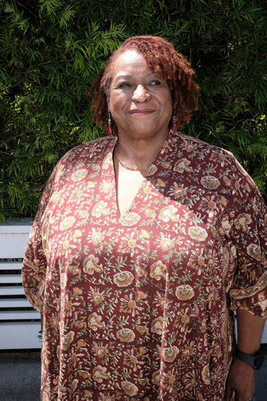 A woman outside standing against greenery wearing a raisin floral tapestry maxi caftan with three quarter balloon sleeves .