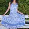 Woman standing in front of white fence and hedge wearing a white with blue tapestry midi dress with a draped neck and  very full skirt.