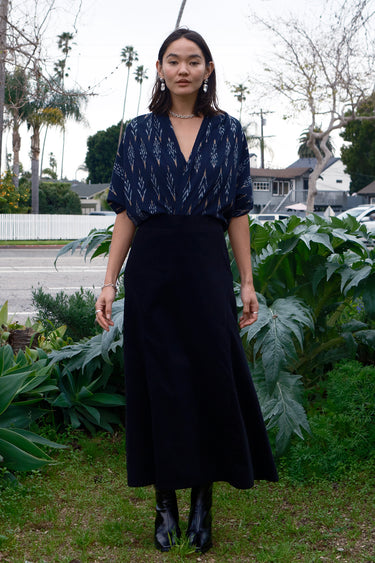 A woman standing outdoors wearing a midi black corduroy skirt with a side zipper, paired with a black top and black boots.