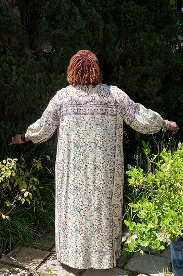 A woman wearing a maxi caftan with a floral pattern in a color that could be described as deep beige ground with tan, green and black paisley tapestry, standing outdoors, back view.